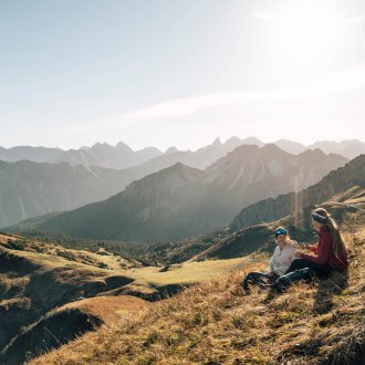 Two people sitting relaxed on a mountain meadow with a view of a stunning mountain range at sunset.