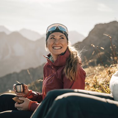 Zwei Frauen sitzen in den Bergen, eine lächelt fröhlich und trinkt aus einer Tasse, während die andere nur teilweise im Bild zu sehen ist.