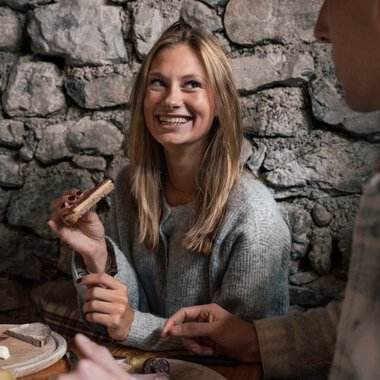 A young blonde woman smiling in a cozy stone setting, talking to a man and holding a small wooden figure. | ©  Kleinwalsertal Tourismus eGen • Photographer: Hannah Bichay