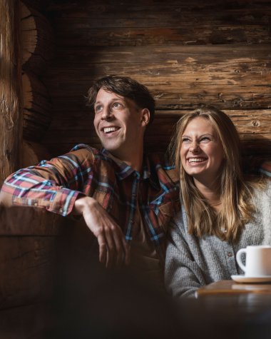 Zwei fröhliche Menschen sitzen in einer gemütlichen Holzhütte, lachen und genießen eine schöne Moment, während sie an einem Tisch mit Tassen sitzen. | © Kleinwalsertal Tourismus eGen • Fotograf: Hannah Bichay