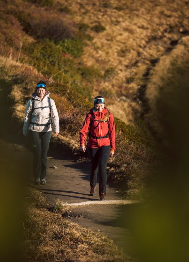 Two women hiking along a trail in a hilly, natural landscape on a sunny day. | ©  Kleinwalsertal Tourismus eGen • Photographer: Hannah Bichay