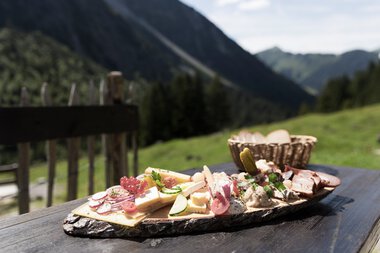 Cheese, sausage, and vegetable platter on a wooden table with mountain scenery in the background, perfect for a mountain hike | © Kleinwalsertal Tourismus eGen • Photographer: Oliver Farys
