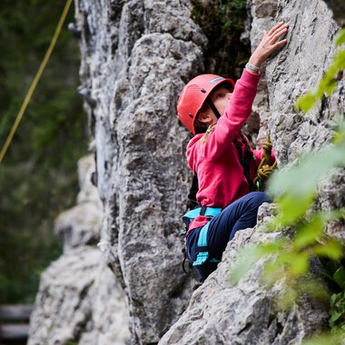 Child climbing in a rock climbing garden, wearing a helmet and harness, outdoors during summer training. | © Kleinwalsertal Tourismus eGen • Photographer: Oliver Farys