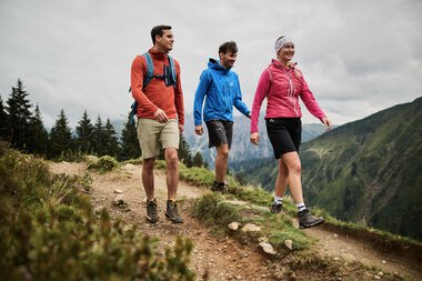Three people hiking on a mountain trail in Kleinwalsertal, surrounded by mountains and fir trees, on a cloudy day. | © Kleinwalsertal Tourismus eGen • Photographer: Oliver Farys