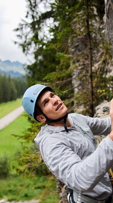Woman climbing at a rock climbing area in the mountains, surrounded by green forests and a path in the background. | © Kleinwalsertal Tourismus eGen • Photographer: Oliver Farys