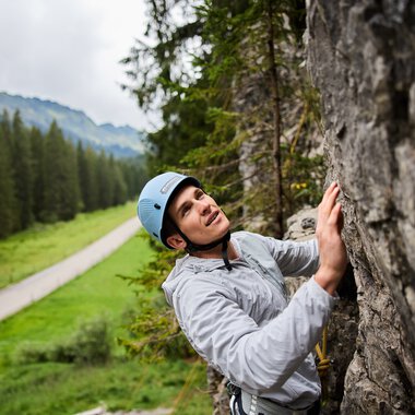 Frau beim Klettern am Klettergarten in den Bergen, umgeben von grünen Wäldern und einer Landschaft mit Weg im Hintergrund. | © Kleinwalsertal Tourismus eGen • Fotograf: Oliver Farys