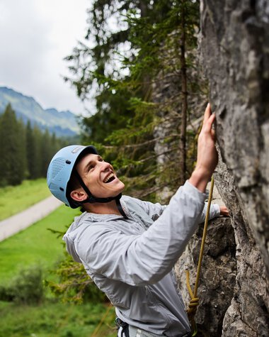 Young woman climbing in a rock climbing area, wearing a helmet and safety gear, with mountains and forests in the background. | © Kleinwalsertal Tourismus eGen • Photographer: Oliver Farys