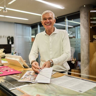 Man at the customer service counter in the office, smiling, with documents and a cash register. | © Kleinwalsertal Tourismus eGen • Photographer: Oliver Farys