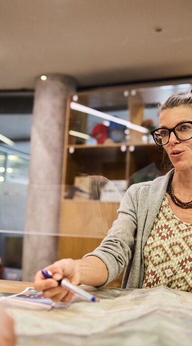 Woman with glasses and curly hair sitting at a desk in a customer service office, speaking friendly, with papers in front of her. | © Kleinwalsertal Tourismus eGen • Photographer: Oliver Farys