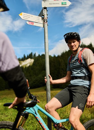 A man taking a break from mountain biking at a signpost in a green, mountainous landscape under a summer sky. | © Kleinwalsertal Tourismus eGen • Photographer: Oliver Farys