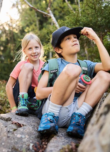 Two children sitting on a rock in a forest, one looking up, the other smiling, surrounded by trees and nature. | © Kleinwalsertal Tourismus eGen • Photographer: Oliver Farys