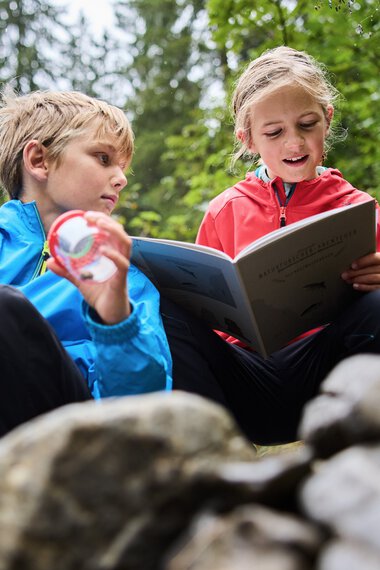 Zwei Kinder sitzen im Wald am Fluss, eines liest aus einem Buch vor, das andere hört aufmerksam zu, umgeben von Bäumen und Natur. | © Kleinwalsertal Tourismus eGen • Fotograf: Oliver Farys