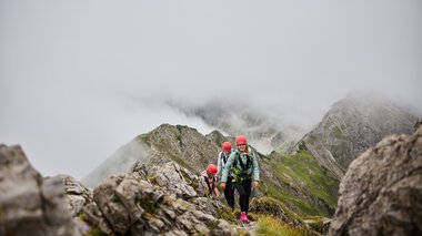 People on a via ferrata in the mountains surrounded by clouds and rocks, with Walser Mountain School logo visible. | © Kleinwalsertal Tourismus eGen • Photographer: Oliver Farys