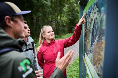 Frau erklärt einer Gruppe von jungen Leuten im Wald eine Informationstafel über die Tier- und Pflanzenwelt. | © Kleinwalsertal Tourismus eGen • Fotograf: Oliver Farys