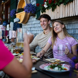 Two smiling people sit outdoors enjoying summer food at a table in front of an alpine hut, with flowers under a window box. | © Kleinwalsertal Tourismus eGen • Photographer: Oliver Farys