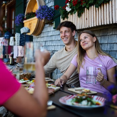 Two smiling people sit outdoors enjoying summer food at a table in front of an alpine hut, with flowers under a window box. | © Kleinwalsertal Tourismus eGen • Photographer: Oliver Farys
