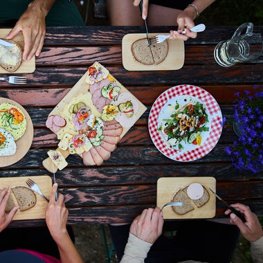 Blick von oben auf einen Tisch mit verschiedenen Käse- und Wurstplatten, Brot, und Salat, umgeben von vier Personen beim Essen. | © Kleinwalsertal Tourismus eGen • Fotograf: Oliver Farys