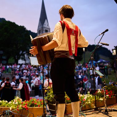 Musician on stage at the Alpenzauber Festival in Kleinwalsertal, playing guitar while the crowd watches, with mountains and a church in the background. | © Kleinwalsertal Tourismus eGen • Photographer: Oliver Farys