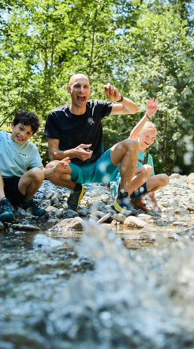 A lively group of four people, including children and an adult, sitting by a creek in a green forest, enjoying playing in the water. | © Kleinwalsertal Tourismus eGen • Photographer: Oliver Farys