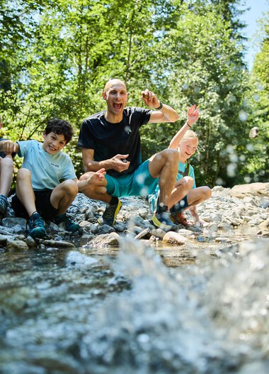 A lively group of four people, including children and an adult, sitting by a creek in a green forest, enjoying playing in the water. | © Kleinwalsertal Tourismus eGen • Photographer: Oliver Farys