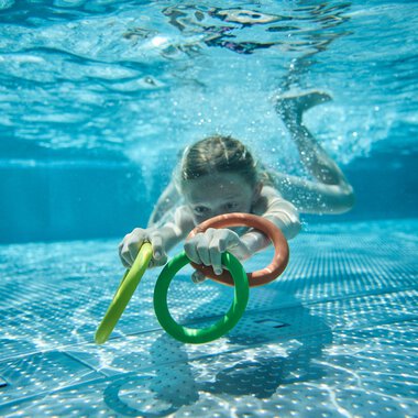 A young girl swimming in an outdoor pool, holding colorful foam noodles and looking forward underwater. | © Kleinwalsertal Tourismus eGen • Photographer: Oliver Farys