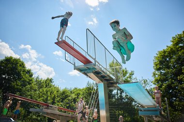Children jumping off a diving board at Freibad Riezlern, surrounded by lush greenery under a sunny sky. | © Kleinwalsertal Tourismus eGen • Photographer: Oliver Farys