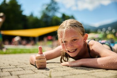 A smiling girl lying on the ground at a swimming pool in Riezlern, giving a thumbs up on a sunny summer day. | © Kleinwalsertal Tourismus eGen • Photographer: Oliver Farys