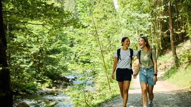 Zwei junge Frauen wandern lachend durch einen grünen Waldweg am Fluss im Sommer 2024, begleitet von Natur und Sonnenschein. | © Kleinwalsertal Tourismus eGen • Fotograf: Oliver Farys