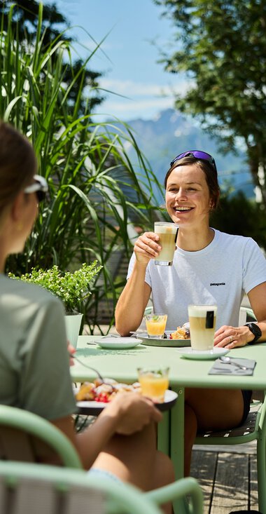 Zwei Frauen genießen eine Mahlzeit und Getränke im Sommer auf einer Terrasse, umgeben von grünen Pflanzen und blauer Himmel. | © Kleinwalsertal Tourismus eGen • Fotograf: Oliver Farys