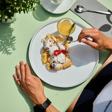 Tisch mit einem Teller mit köstlichem Kaiserschmarrn, garnierte mit Puderzucker und Kirschen, daneben ein Glas frisch gepresster Orangensaft. | © Kleinwalsertal Tourismus eGen • Fotograf: Oliver Farys