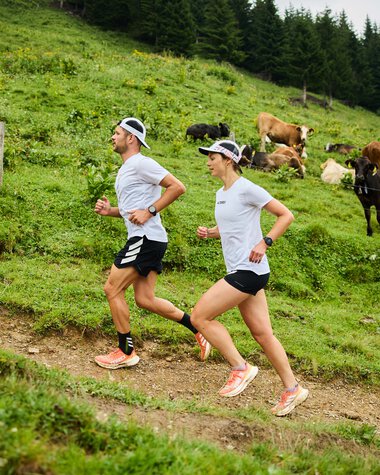 Zwei Frauen joggen einen grasbewachsenen Trail im Grünen, umgeben von Kühen und Blick auf den Wald im Hintergrund. | © Kleinwalsertal Tourismus eGen • Fotograf: Oliver Farys