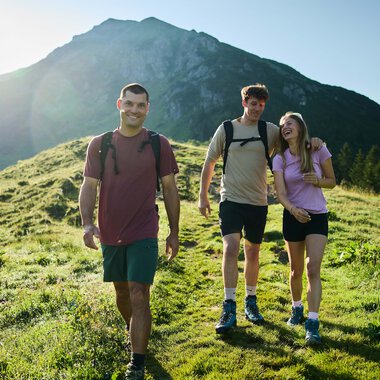 Drei junge Erwachsene wandern fröhlich auf einer grünen Wiese mit Bergen im Hintergrund bei Sonnenschein. | © Kleinwalsertal Tourismus eGen • Fotograf: Oliver Farys