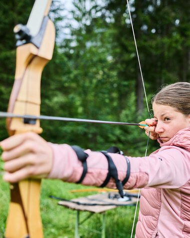 Girl practicing archery at the wilderness school in Kleinwalsertal, surrounded by green nature. | © Kleinwalsertal Tourismus eGen • Photographer: Oliver Farys