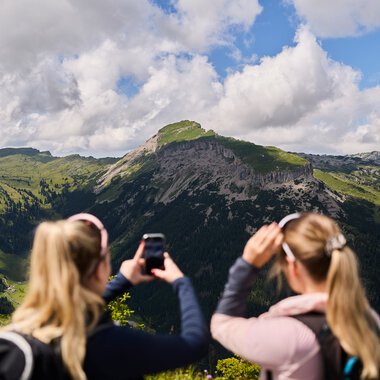 Two women taking photos of a stunning mountain landscape in Kleinwalsertal, surrounded by forests and cloudy sky. | © Kleinwalsertal Tourismus eGen • Photographer: Oliver Farys