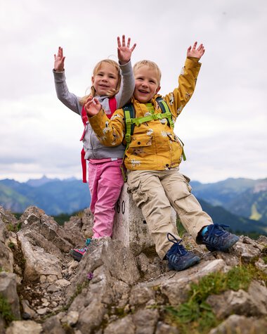 Two happy children on a mountain hike, sitting on a rock with backpacks, raising their hands in the air, smiling during summer. | © Kleinwalsertal Tourismus eGen • Photographer: Oliver Farys