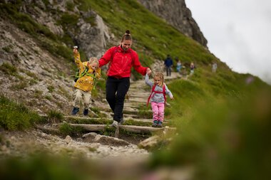 Mother and two children hiking on a mountain trail in Kleinwalsertal, surrounded by lush greenery on a sunny day. | © Kleinwalsertal Tourismus eGen • Photographer: Oliver Farys