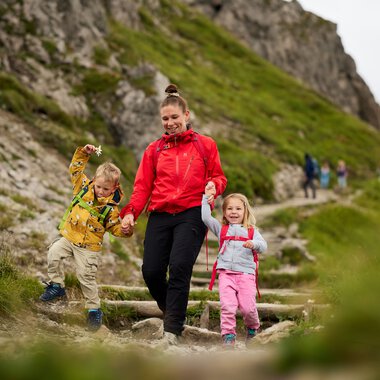 Family hiking in the mountains on a trail, children and adults with green mountain slopes in the background. | © Kleinwalsertal Tourismus eGen • Photographer: Oliver Farys