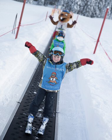 A child skiing happily on a snow track, with a large smiling mascot in the background during a winter ski trip. | © Kleinwalsertal Tourismus eGen • Photographer: Oliver Farys