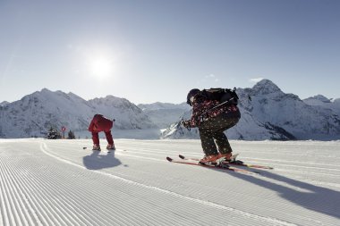 Zwei Skifahrer auf frisch präpariertem Schneeboden in den Bergen, Sonne und schneebedeigte Gipfel im Hintergrund. | © Kleinwalsertal Tourismus eGen • Fotograf: Oliver Farys