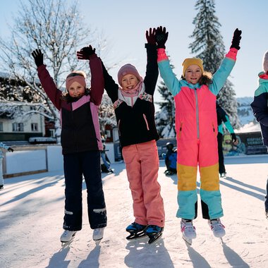Children ice skating on a frozen lake in winter, joyfully raising their arms, with snow-covered trees and other skaters around. | © Kleinwalsertal Tourismus eGen • Photographer: Oliver Farys