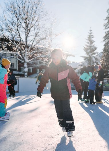 Kinder beim Eislaufen auf einem Eisparkplatz im Winter, umgeben von Bäumen und Gebäuden, sonniger Tag. | © Kleinwalsertal Tourismus eGen • Fotograf: Oliver Farys