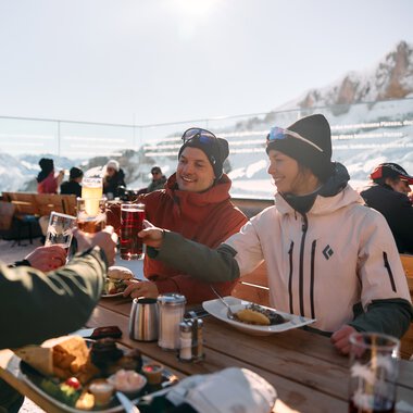 Three friends having lunch outdoors with a mountain view in Kleinwalsertal, toasting with drinks. | © Kleinwalsertal Tourismus eGen • Photographer: Oliver Farys