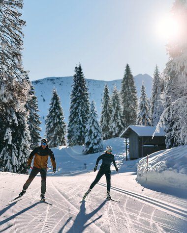 Two cross-country skiers enjoying skiing in a snowy winter forest in Kleinwalsertal under sunny skies. | © Kleinwalsertal Tourismus eGen • Photographer: Oliver Farys