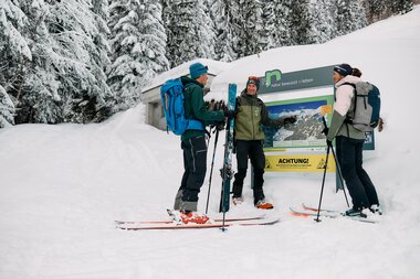 Three cross-country skiers in front of an informational sign in a snowy forest near Kleinwalsertal, winter 2024. | © Kleinwalsertal Tourismus eGen • Photographer: Oliver Farys