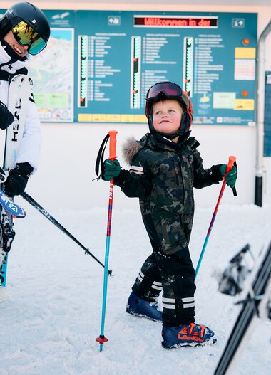 Children with ski gear at the ski resort, a display board in the background shows lift info. Snowy winter scene with ski equipment. | © Kleinwalsertal Tourismus eGen • Photographer: Oliver Farys