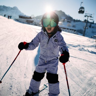 Kleines Kind beim Skifahren auf einer Piste im Snowboard- und Skigebiet im Kleinwalsertal, umgeben von Bergen, mit Skiern und -stöcken, trägt Skibrille und Winterkleidung. | © Kleinwalsertal Tourismus eGen • Fotograf: Oliver Farys