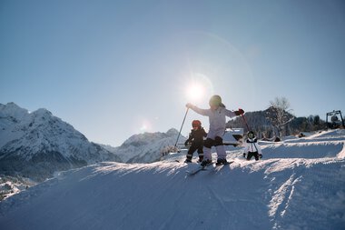 Kinder beim Skifahren auf einer schneebedeckten Piste in den Bergen bei Sonnenlicht, begleitet von einem Erwachsenen. | © Kleinwalsertal Tourismus eGen • Fotograf: Oliver Farys