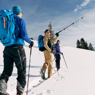 Three skitourers with backpacks in a snowy landscape, one taking a selfie-like photo during a winter tour in Kleinwalsertal. | © Kleinwalsertal Tourismus eGen • Photographer: Oliver Farys