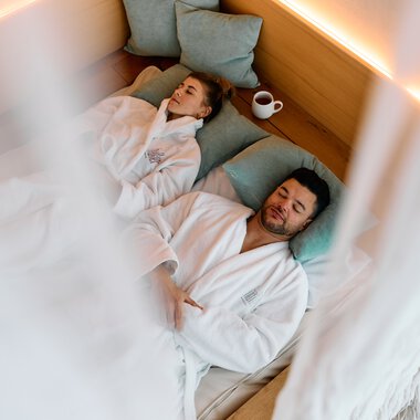 A man and a woman are lying on lounge beds in a wellness spa area, wrapped in bathrobes, with pillows and a cup. They look relaxed and calm. | © Kleinwalsertal Tourismus eGen • Photographer: Oliver Farys