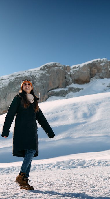 Two people winter hiking in a snowy mountain landscape with rocks in the background and a sunny sky. | © Kleinwalsertal Tourismus eGen • Photographer: Oliver Farys